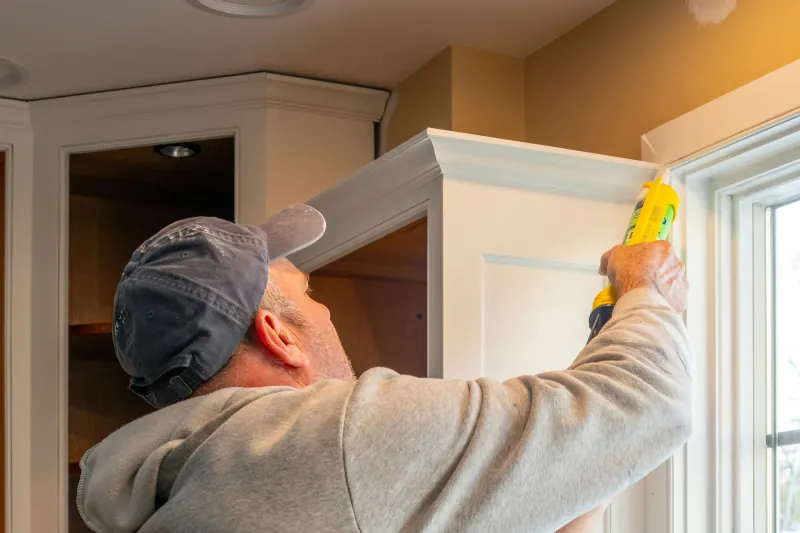 Photo of a man using a caulk gun to seal trim on a white upper cabinet beside open cabinet boxes.
