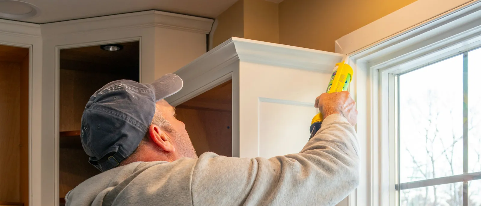 Photo of a man using a caulk gun to seal trim on a white upper cabinet beside open cabinet boxes.