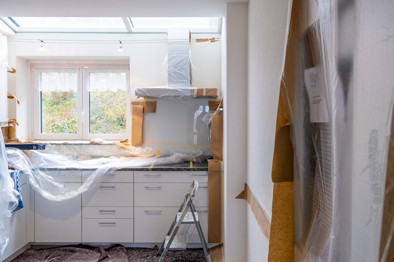 A kitchen mid-renovation, cabinets and counters masked with plastic sheeting and paper, a stepladder in front of the run