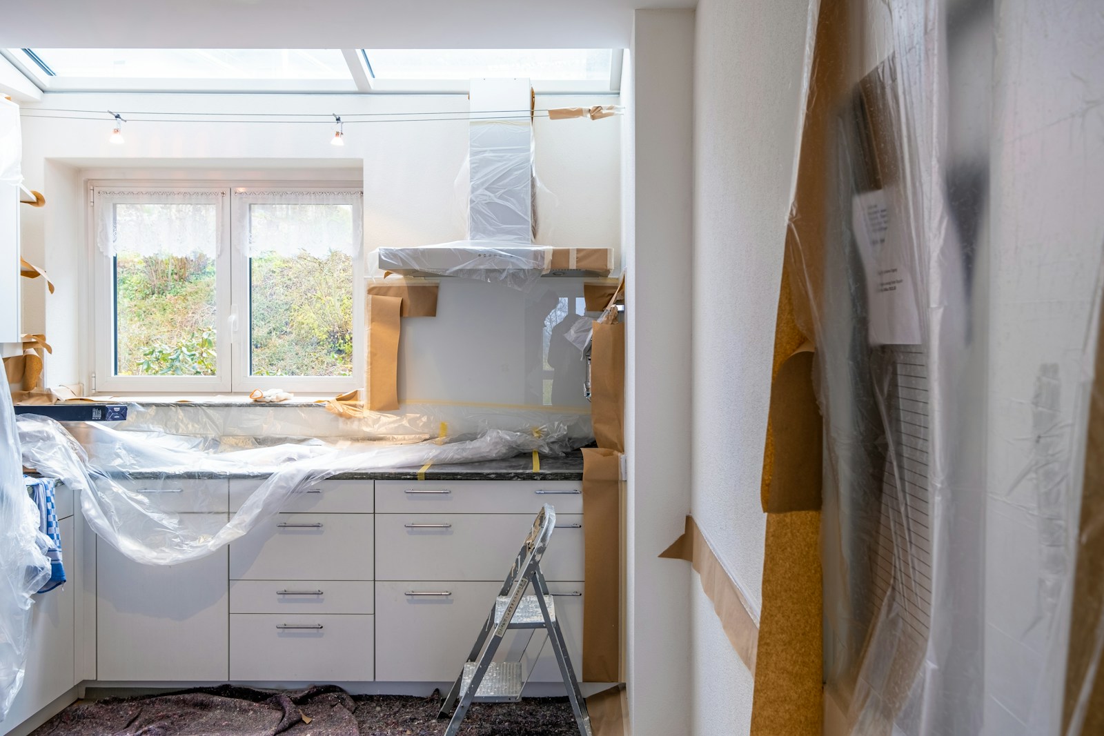 A kitchen mid-renovation, cabinets and counters masked with plastic sheeting and paper, a stepladder in front of the run