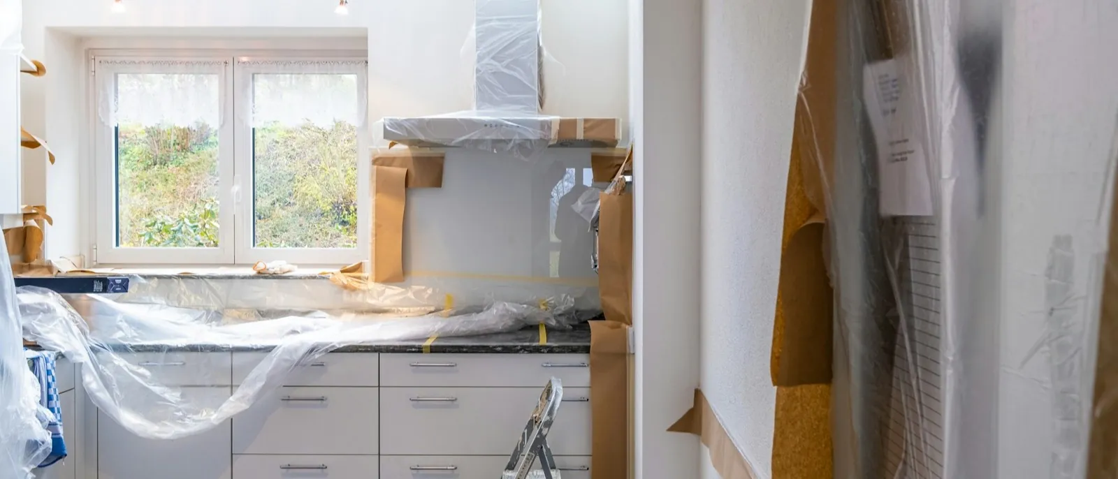 A kitchen mid-renovation, cabinets and counters masked with plastic sheeting and paper, a stepladder in front of the run