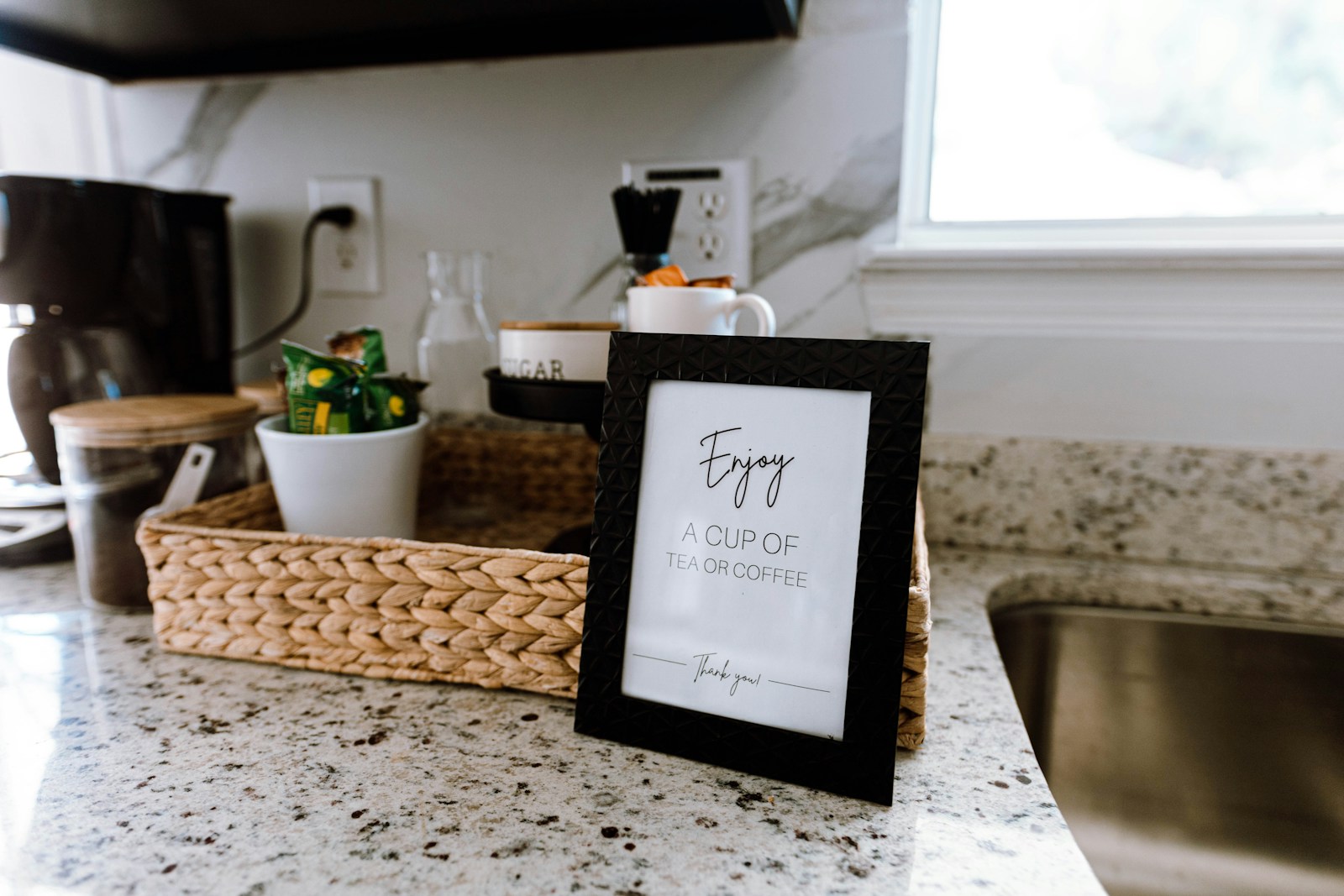 A countertop coffee station arranged in a woven tray with mugs, canisters, and a framed sign