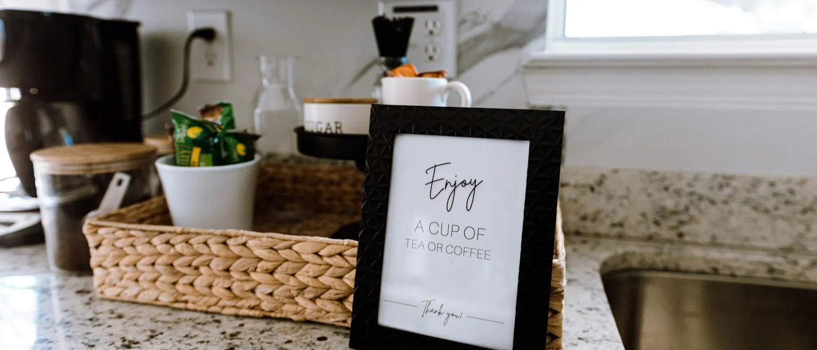 A countertop coffee station arranged in a woven tray with mugs, canisters, and a framed sign