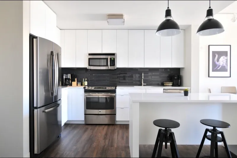 White flat-front modular kitchen with a dark tile backsplash, stainless appliances, and black adjustable stools at a white island