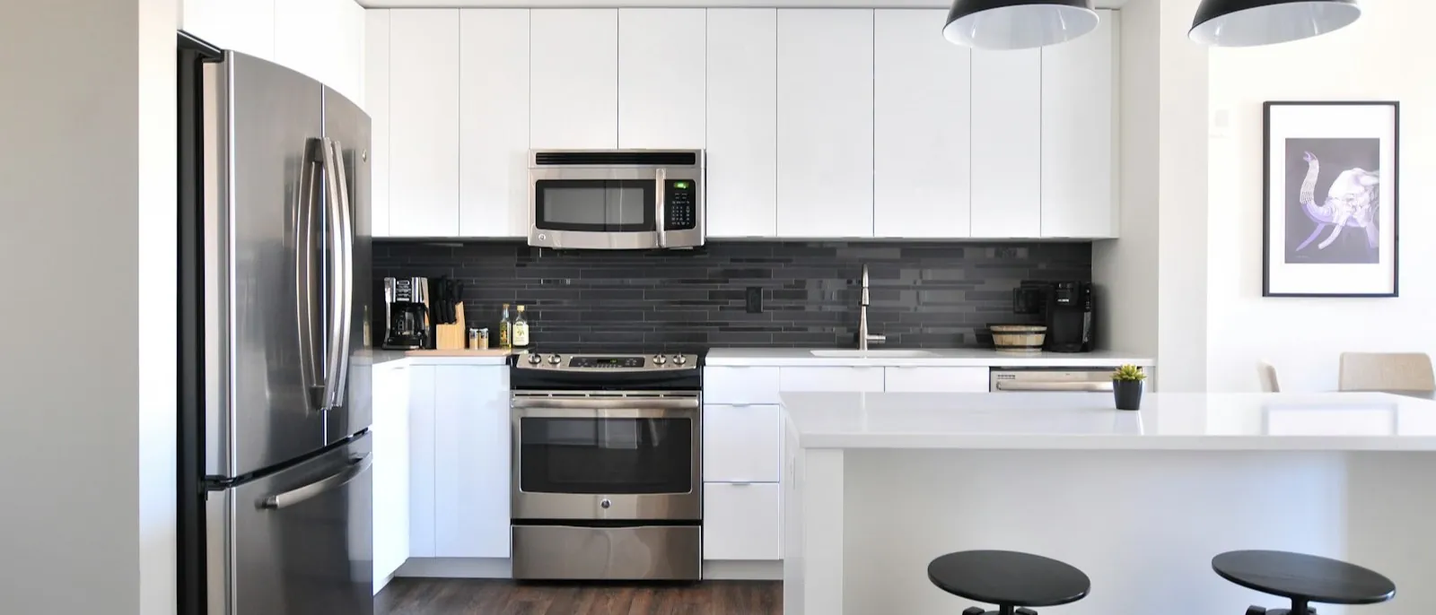 White flat-front modular kitchen with a dark tile backsplash, stainless appliances, and black adjustable stools at a white island