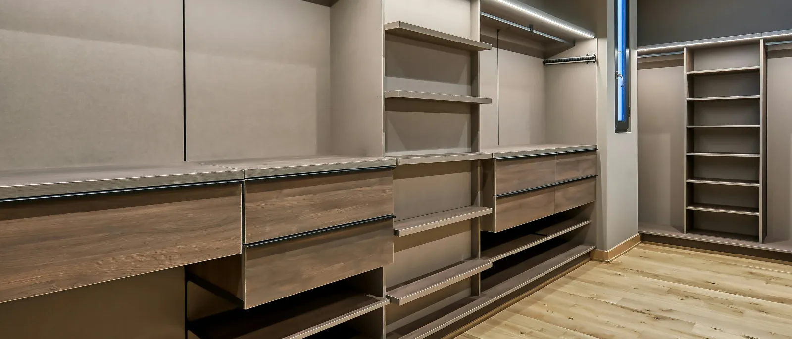 Photo of an empty walk-in closet system with wood-grain drawers, open shelves, a shelf tower and strip lights above a light wood floor.