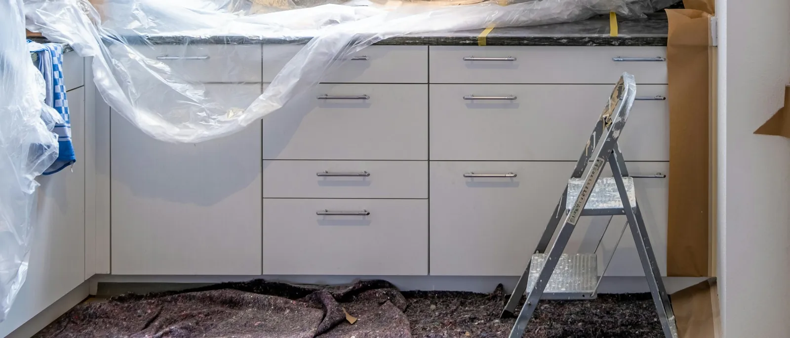 Photo of new white base cabinets during a kitchen installation, with plastic sheeting over the countertop and a step ladder in front.