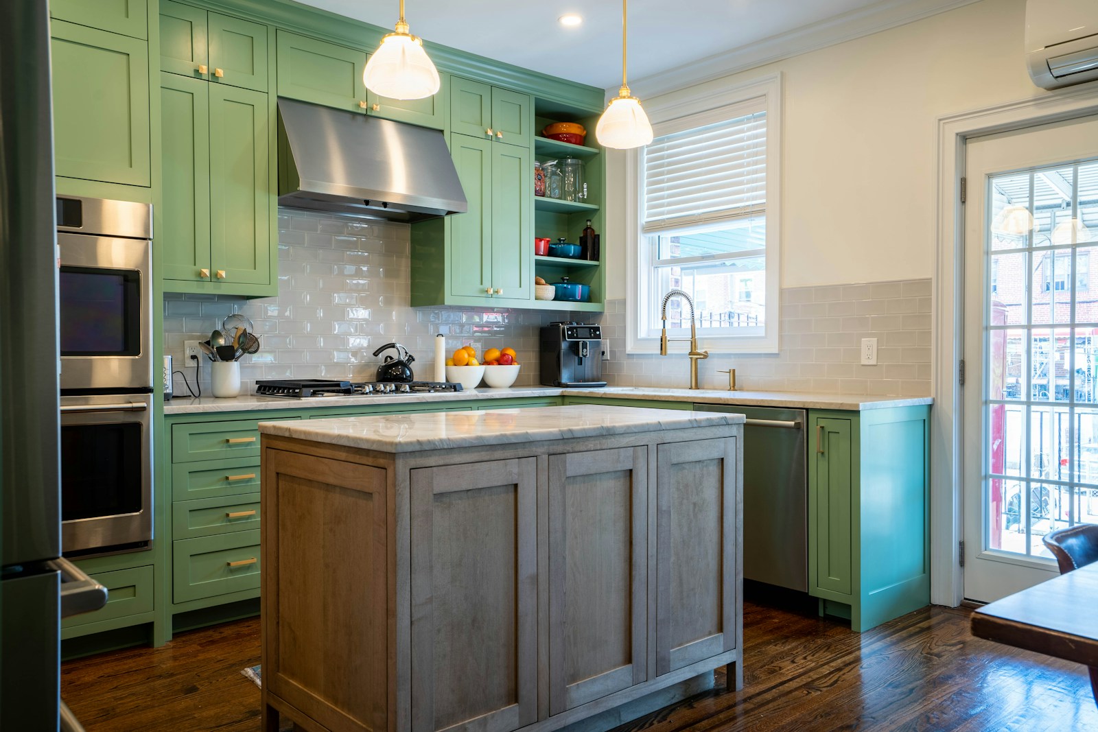 A kitchen with green shaker cabinets, brass hardware, and a wood island under pendant lights