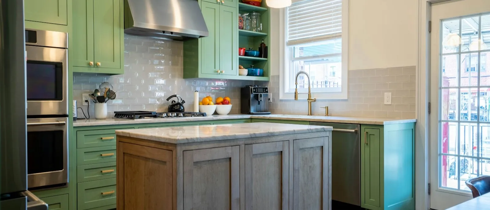 A kitchen with green shaker cabinets, brass hardware, and a wood island under pendant lights